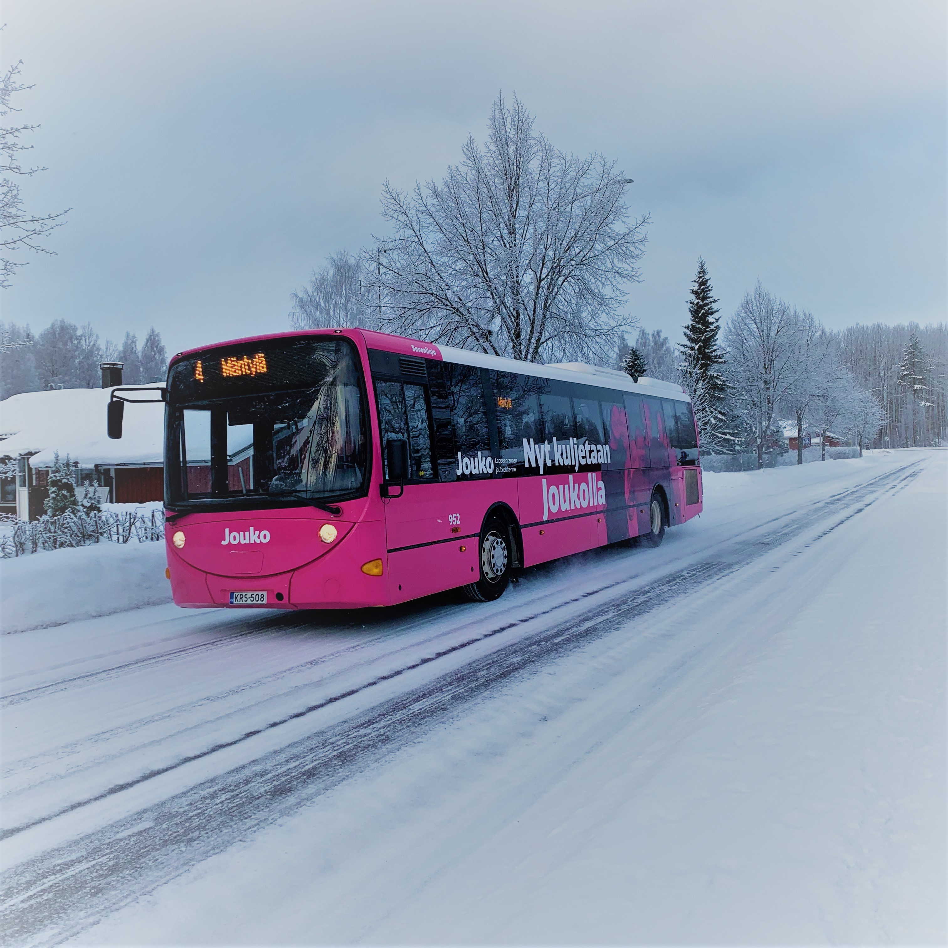 Pinkki bussi talvisella, lumisella tiellä. Takana talo ja puita. Bussissa lukee ”4 Mäntylä” ”Jouko” ja ”Nyt kuljetaan Joukolla”.